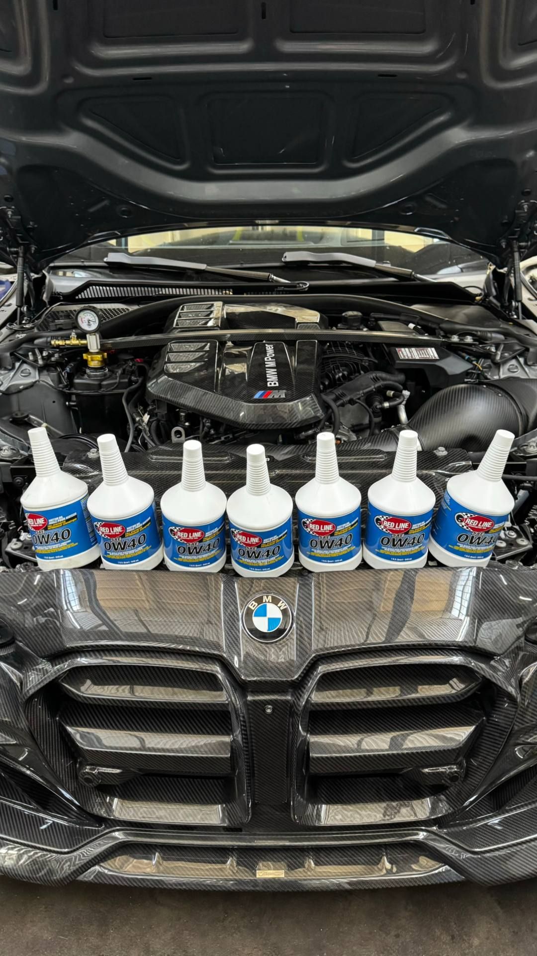 Șapte sticle Red Line 0W40 pe grila unui BMW M cu capota deschisă și motorul M Power