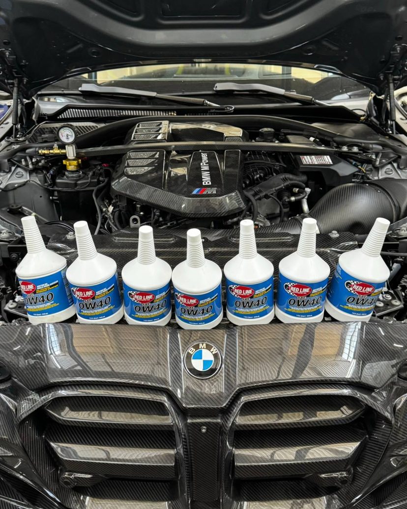 Șapte sticle Red Line 0W40 pe grila unui BMW M cu capota deschisă și motorul M Power