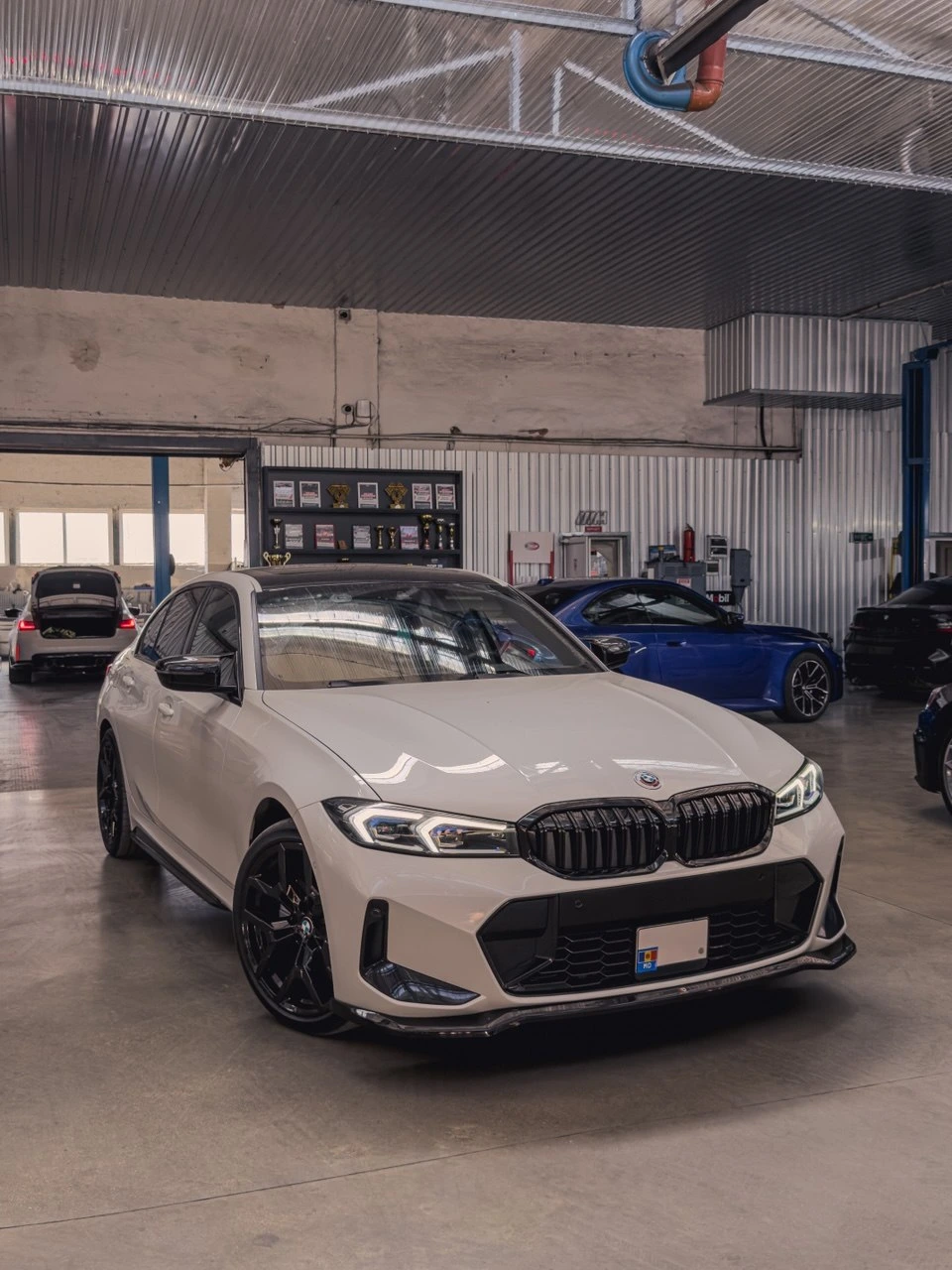 BMW Seria 3 G20 albă în atelier, în spate un raft cu cupe