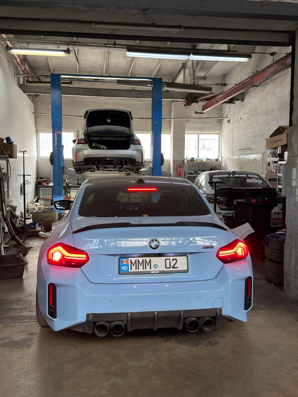 BMW M2 bleu văzută din spate în atelier, deasupra — un BMW pe elevator
